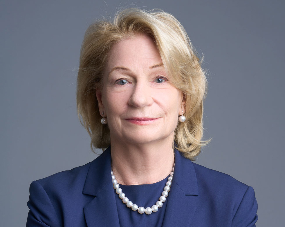 Marilyn Simons wearing a navy blue suit and wearing pearls with her arms crossed.