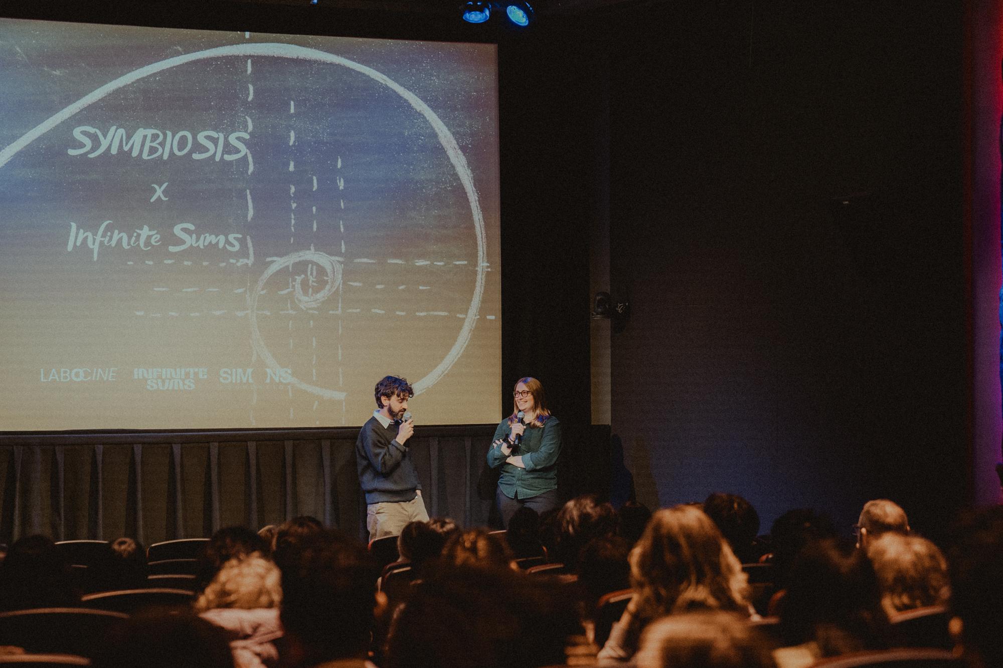 A photo of Sonya Hansen and Diego Murillo at the Symbiosis film screening, standing in front of a screen that says Symbiosis x Infinite Sums.