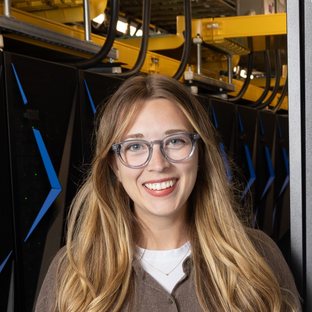 A woman with long blonde hair and glasses smiles, standing in front of server racks with blue lights and yellow cables in a data center.