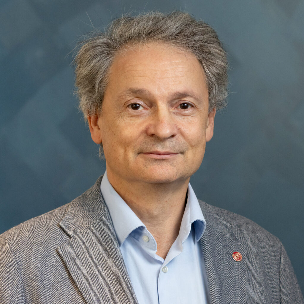 A middle-aged man with gray hair wearing a light blue button-down shirt and a patterned blazer.