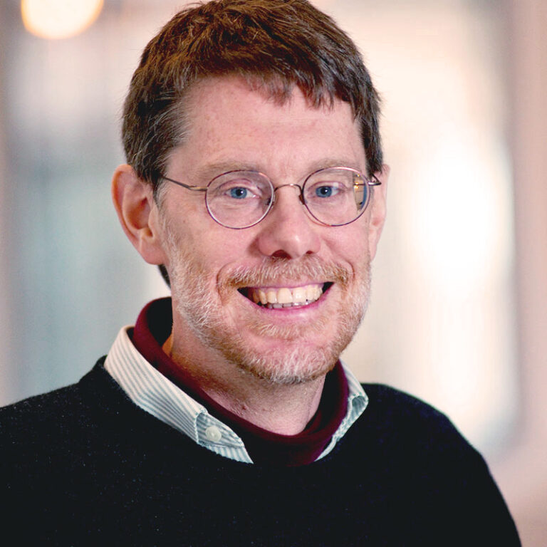 A smiling man with short brown hair and round glasses, wearing a dark sweater over a collared shirt.