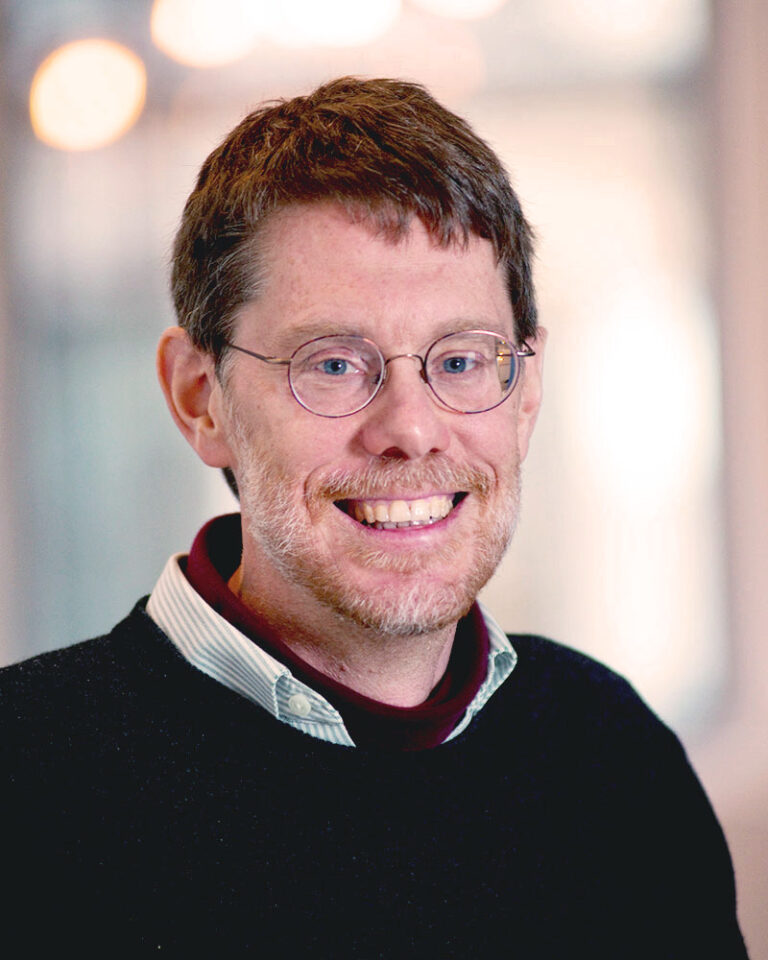 A smiling man with short brown hair and round glasses, wearing a dark sweater over a collared shirt.