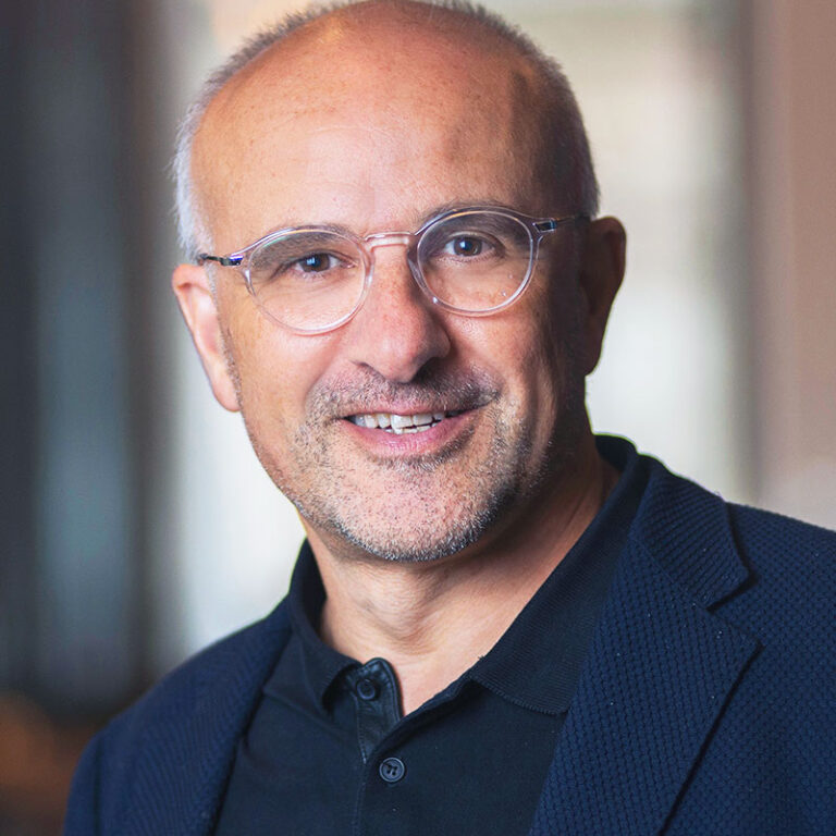 A middle-aged man with short hair and glasses wearing a dark polo shirt and blazer smiles at the camera.