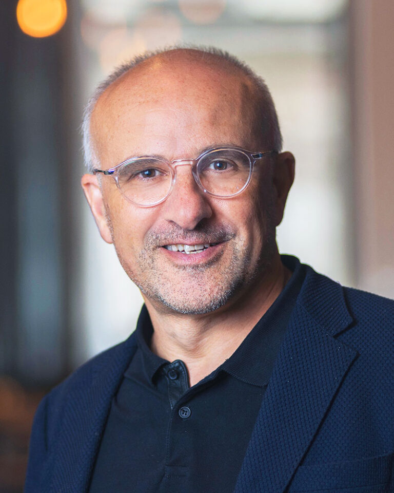 A middle-aged man with short hair and glasses wearing a dark polo shirt and blazer smiles at the camera.