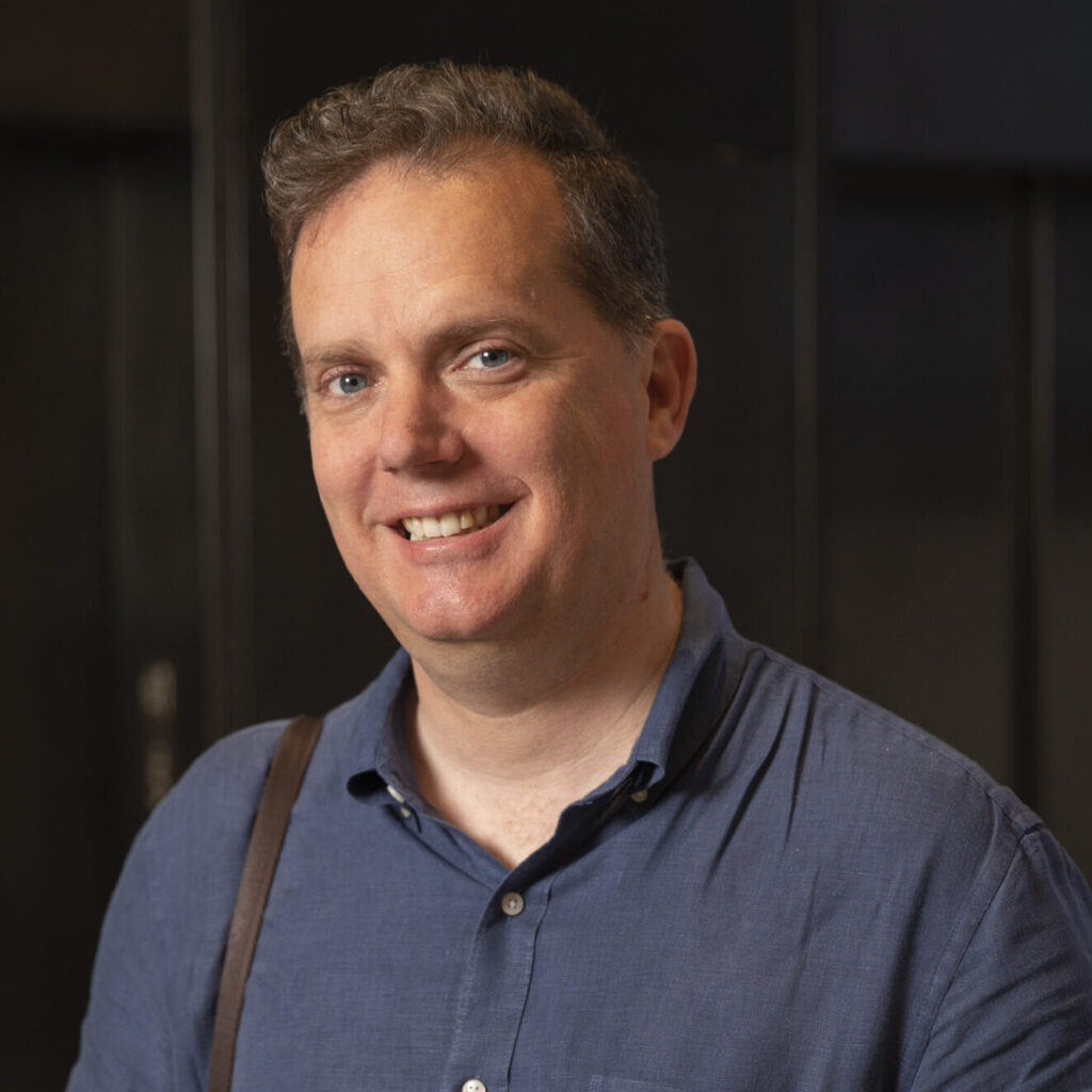 A man wearing a blue short-sleeve shirt smiles at the camera. He has short hair and carries a brown strap over his shoulder. The background is dark and out of focus.