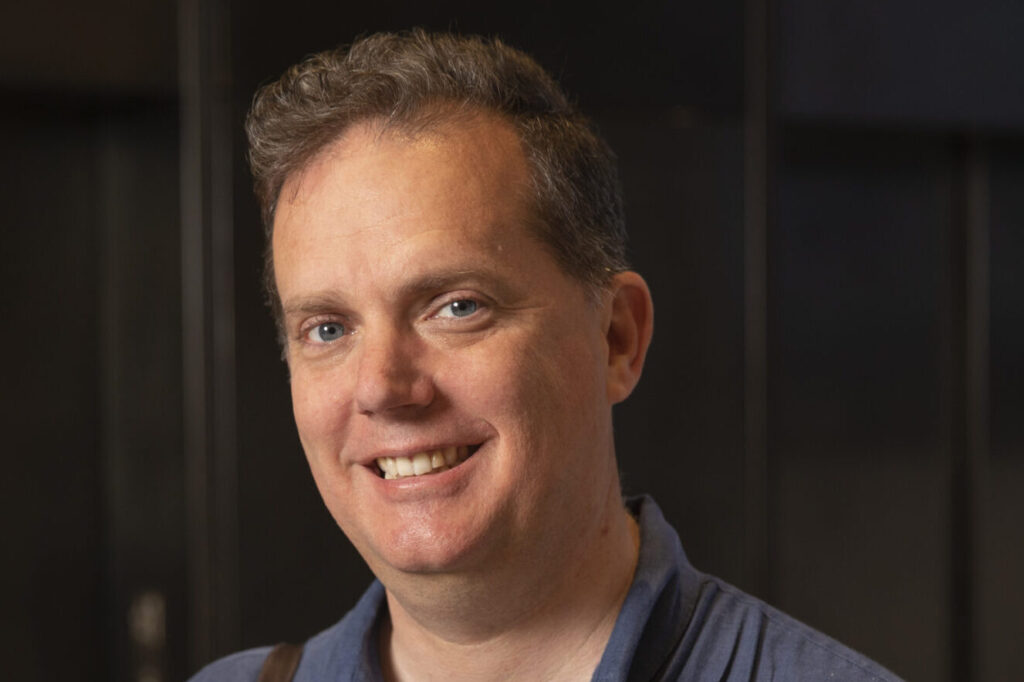 A man wearing a blue short-sleeve shirt smiles at the camera. He has short hair and carries a brown strap over his shoulder. The background is dark and out of focus.