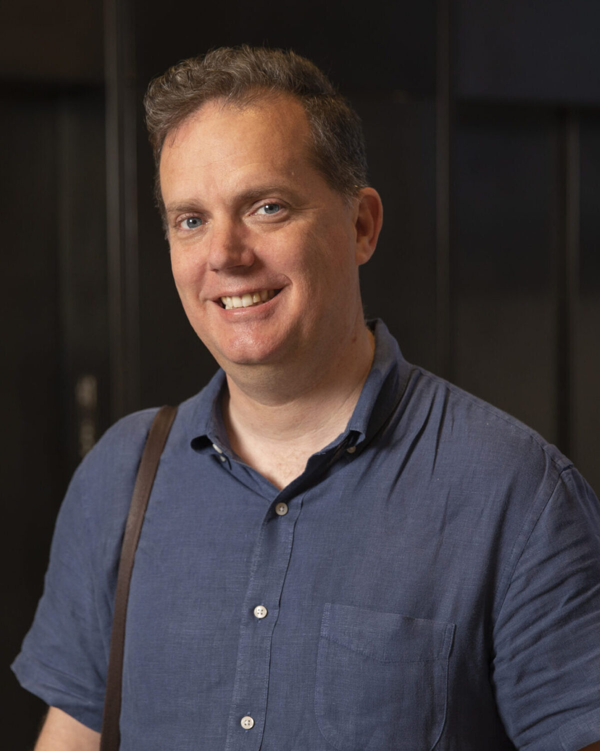 A man wearing a blue short-sleeve shirt smiles at the camera. He has short hair and carries a brown strap over his shoulder. The background is dark and out of focus.