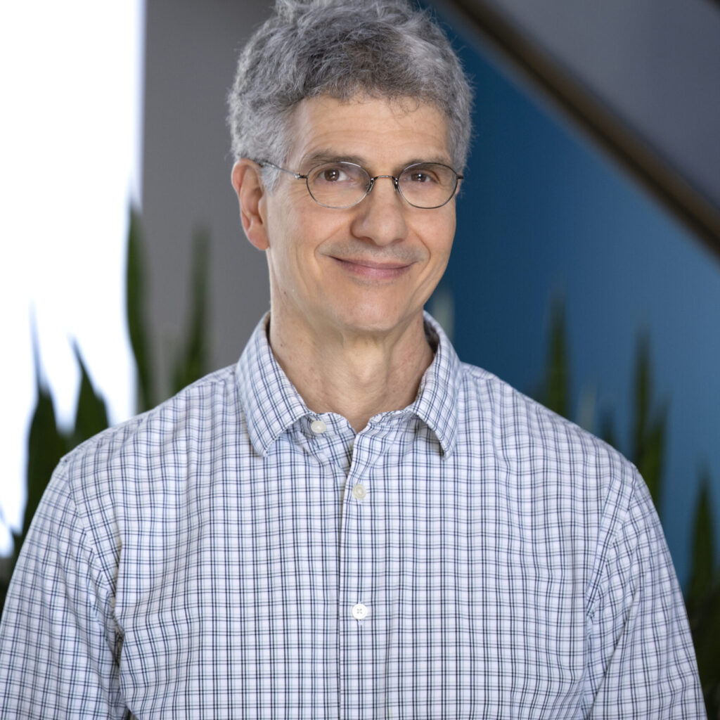 A middle-aged man with gray hair and glasses wearing a light-colored plaid button-down shirt.