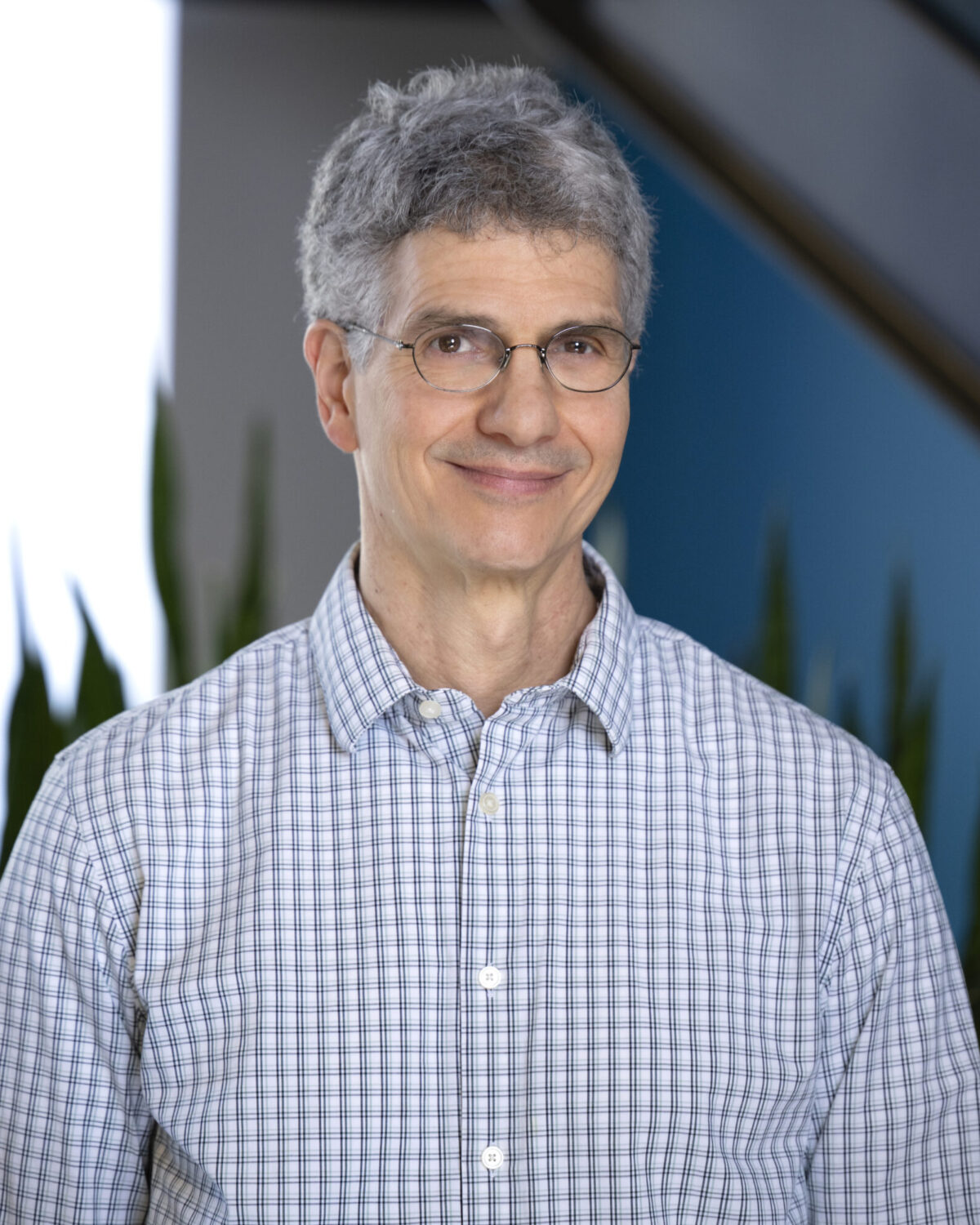 A middle-aged man with gray hair and glasses wearing a light-colored plaid button-down shirt.