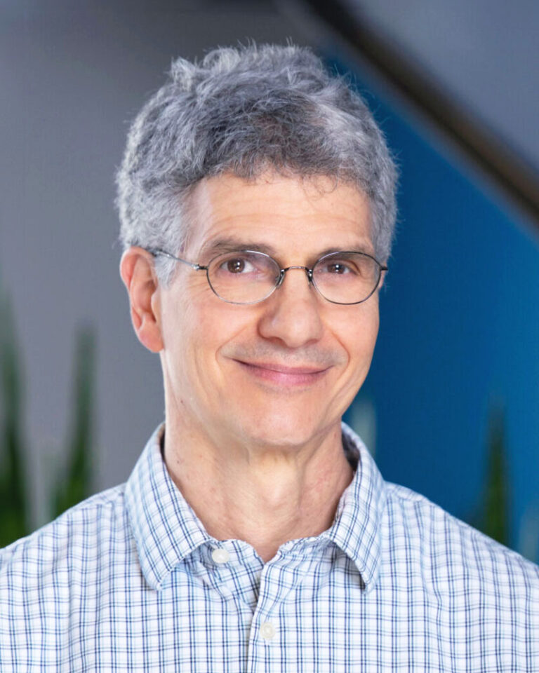 Portrait of a smiling man with graying hair wearing wire-rimmed glasses and a light-colored plaid button-down shirt.