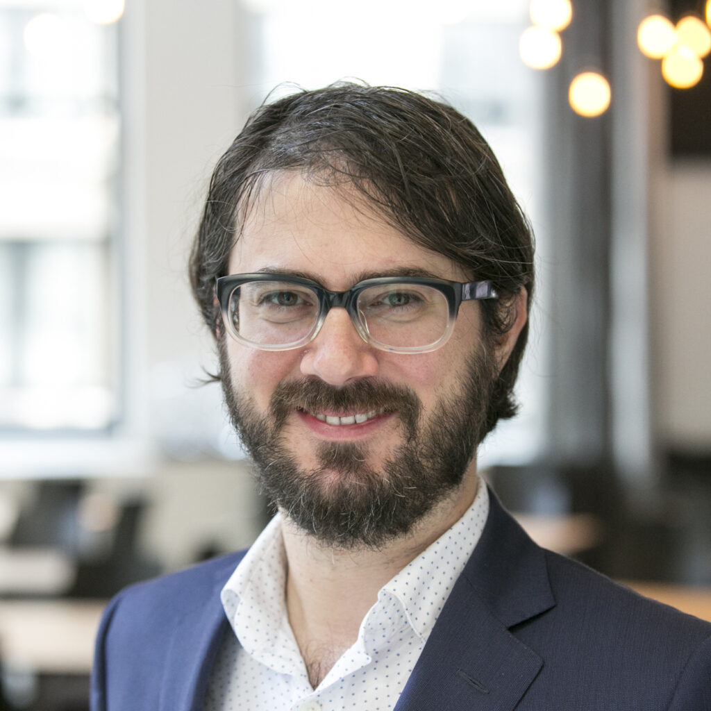 A man with glasses and a beard, wearing a navy blazer and white shirt, smiles in a bright, modern office with blurred lights in the background.