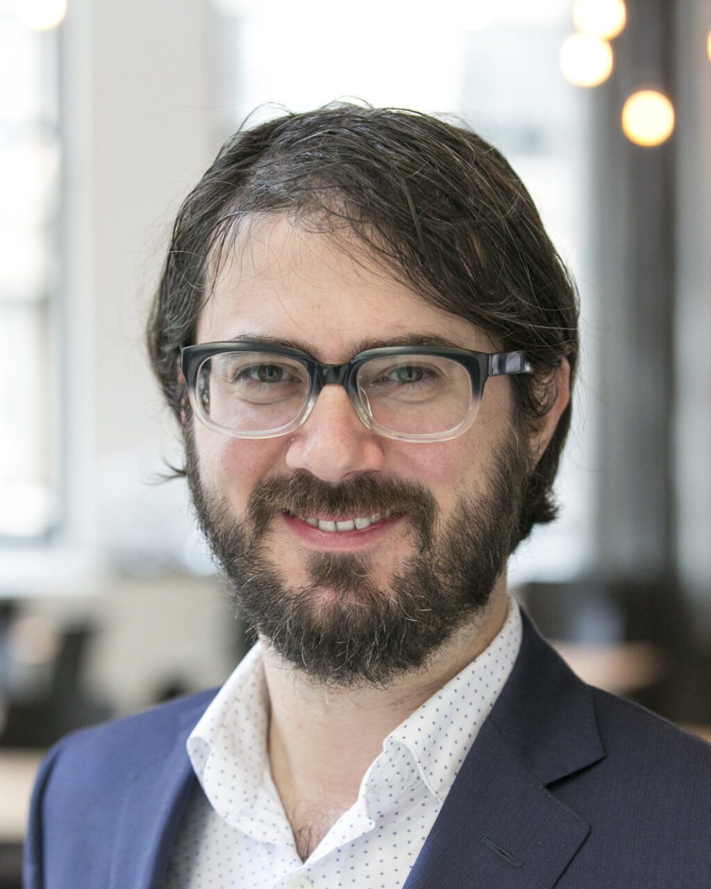 A man with glasses and a beard, wearing a navy blazer and white shirt, smiles in a bright, modern office with blurred lights in the background.