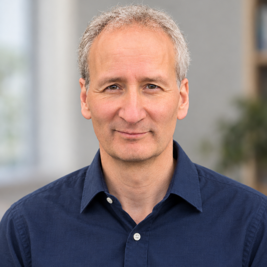 A middle-aged man with short gray hair, wearing a dark blue button-up shirt, is looking at the camera with a neutral expression. The background is blurred indoors with hints of greenery.