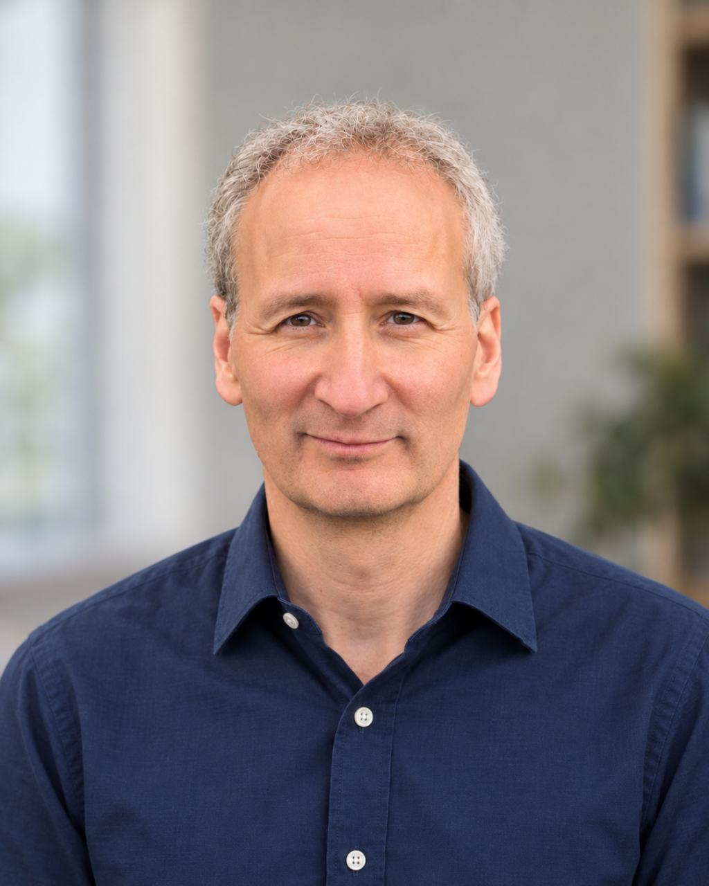 A middle-aged man with short gray hair, wearing a dark blue button-up shirt, is looking at the camera with a neutral expression. The background is blurred indoors with hints of greenery.
