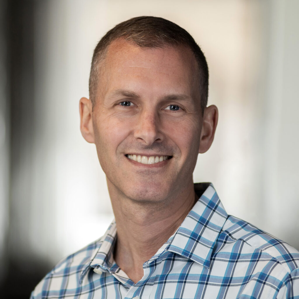 A man with short hair smiles at the camera. He is wearing a blue and white plaid shirt, and the background is softly blurred, keeping the focus on his face.