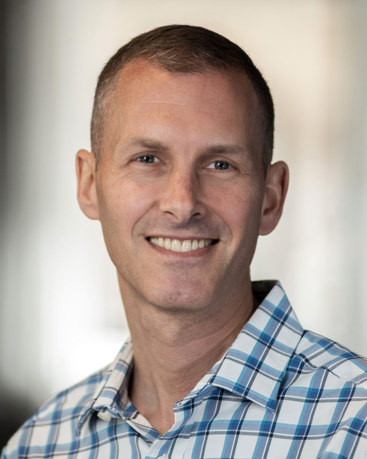 A man with short hair smiles at the camera. He is wearing a blue and white plaid shirt, and the background is softly blurred, keeping the focus on his face.