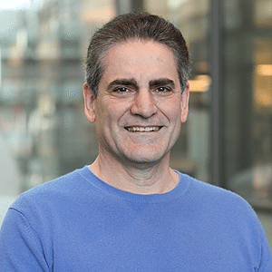 A middle-aged man with short dark hair, wearing a blue sweater, smiles at the camera while standing in front of a blurred glass building background.