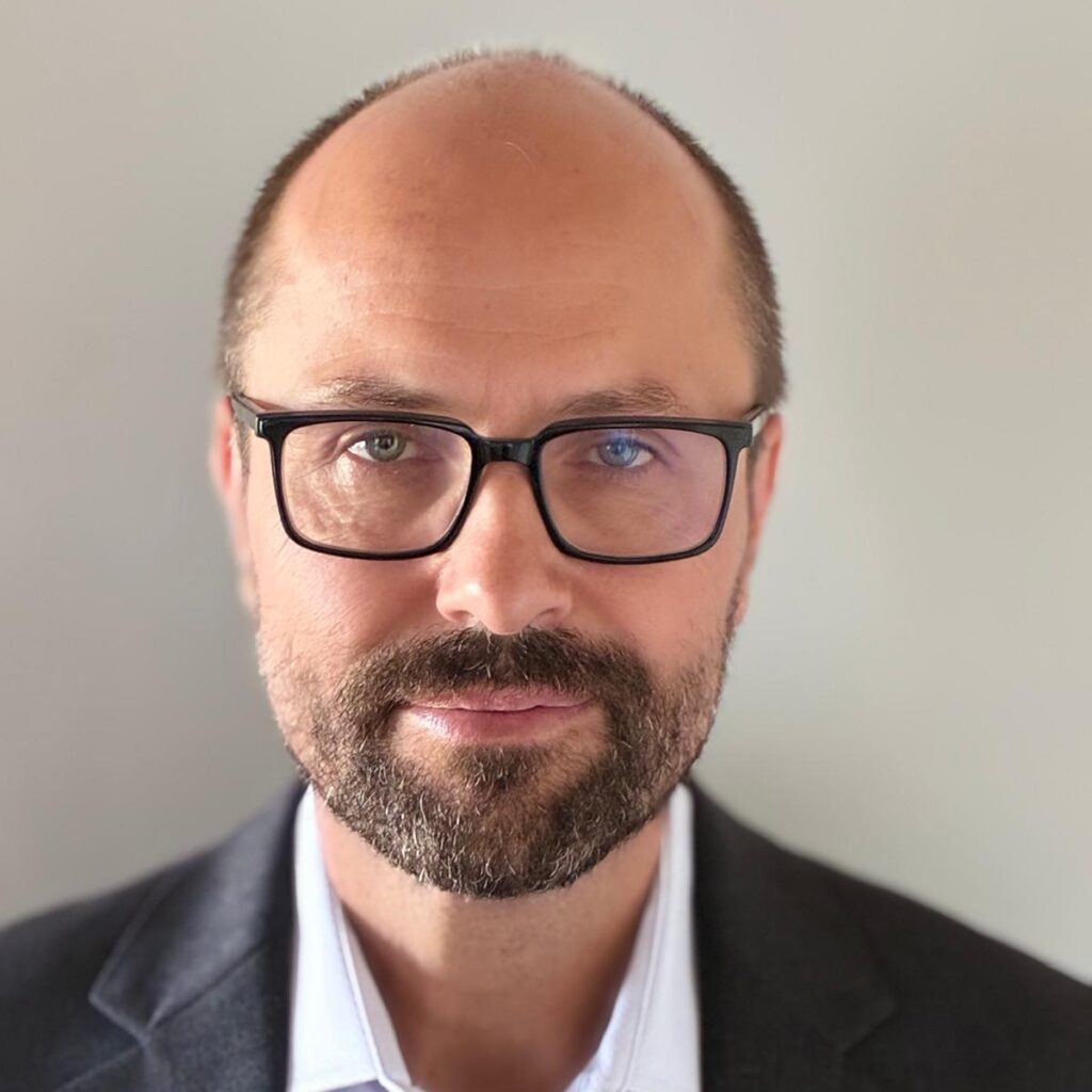 A man with glasses, a short beard, and a receding hairline is wearing a dark blazer and light-colored shirt, facing the camera against a plain background.