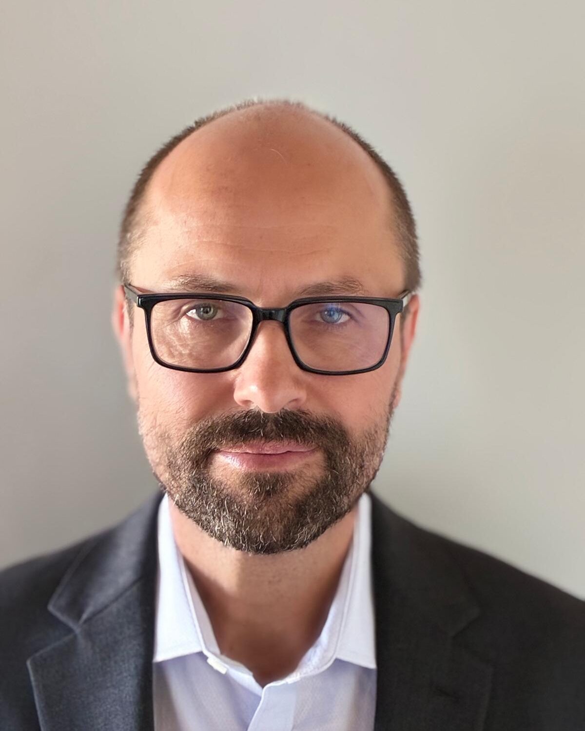 A man with glasses, a short beard, and a receding hairline is wearing a dark blazer and light-colored shirt, facing the camera against a plain background.