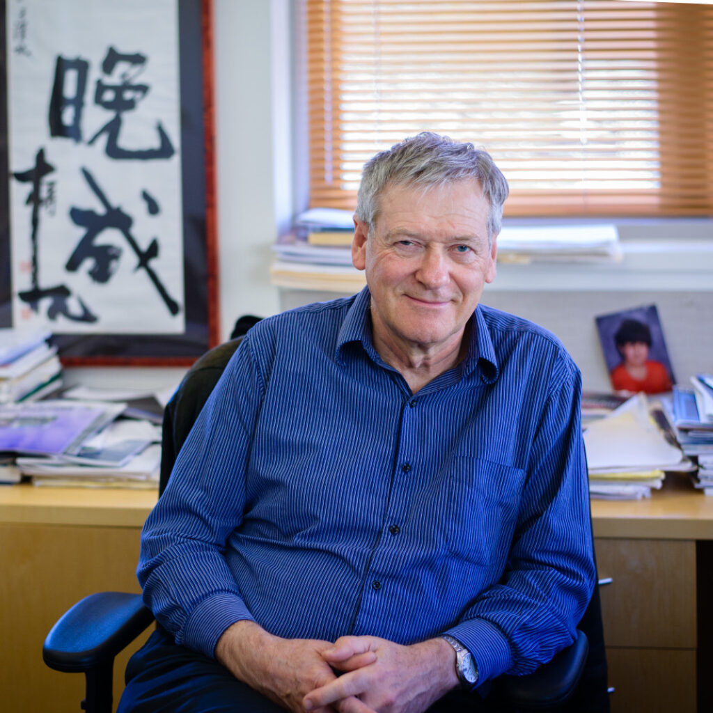 An older man in a blue shirt sits smiling in an office with stacks of papers, a framed calligraphy piece, and a photo of a child on the desk behind him.