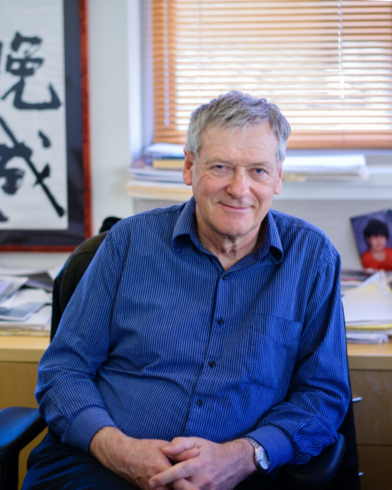 An older man in a blue shirt sits smiling in an office with stacks of papers, a framed calligraphy piece, and a photo of a child on the desk behind him.