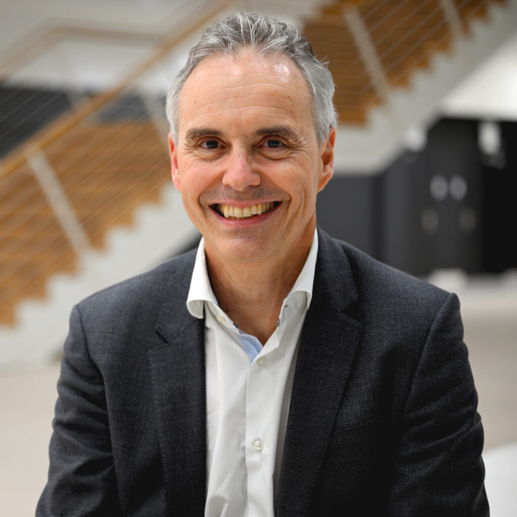 A smiling middle-aged man in a suit sits indoors with a modern wooden staircase and blurred background behind him.