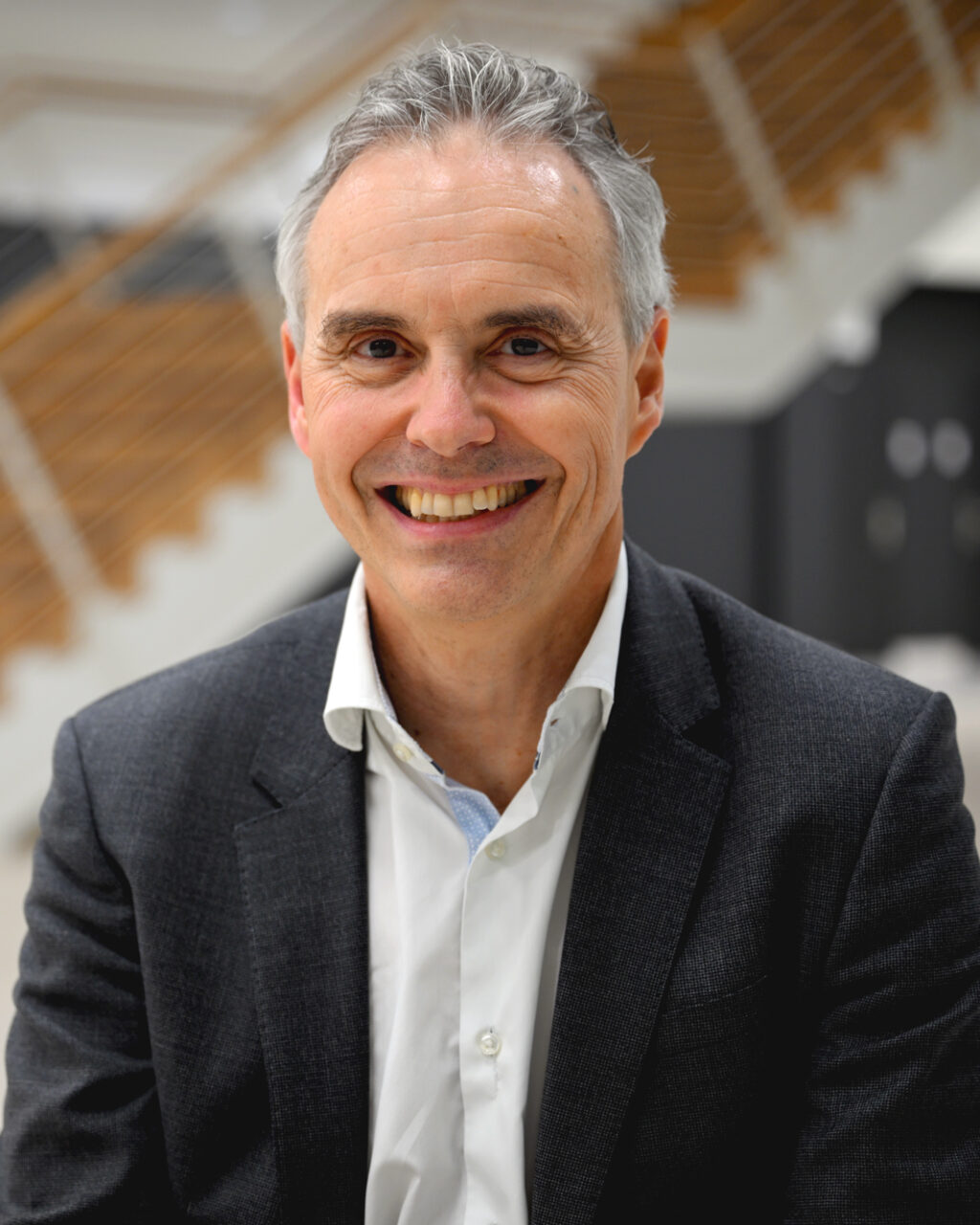 A smiling middle-aged man in a suit sits indoors with a modern wooden staircase and blurred background behind him.