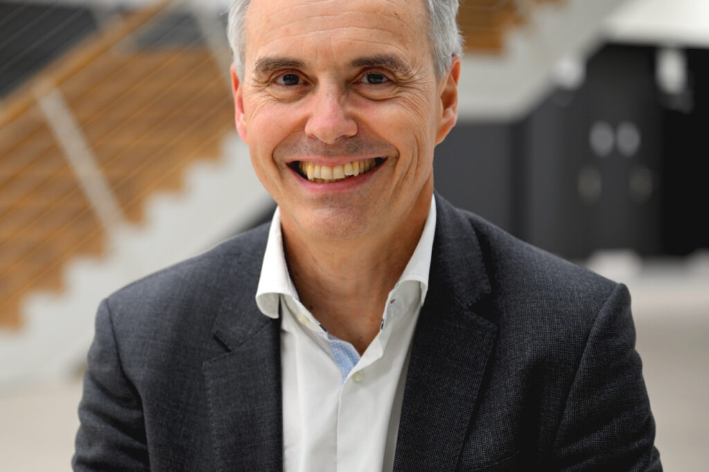 A smiling middle-aged man in a suit sits indoors with a modern wooden staircase and blurred background behind him.