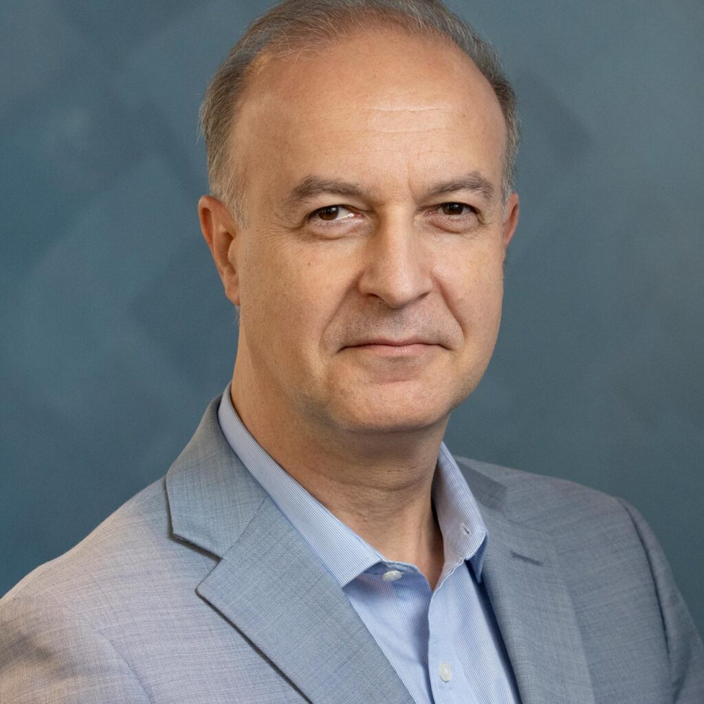 A middle-aged man with short hair wearing a light blue dress shirt and a grey blazer.