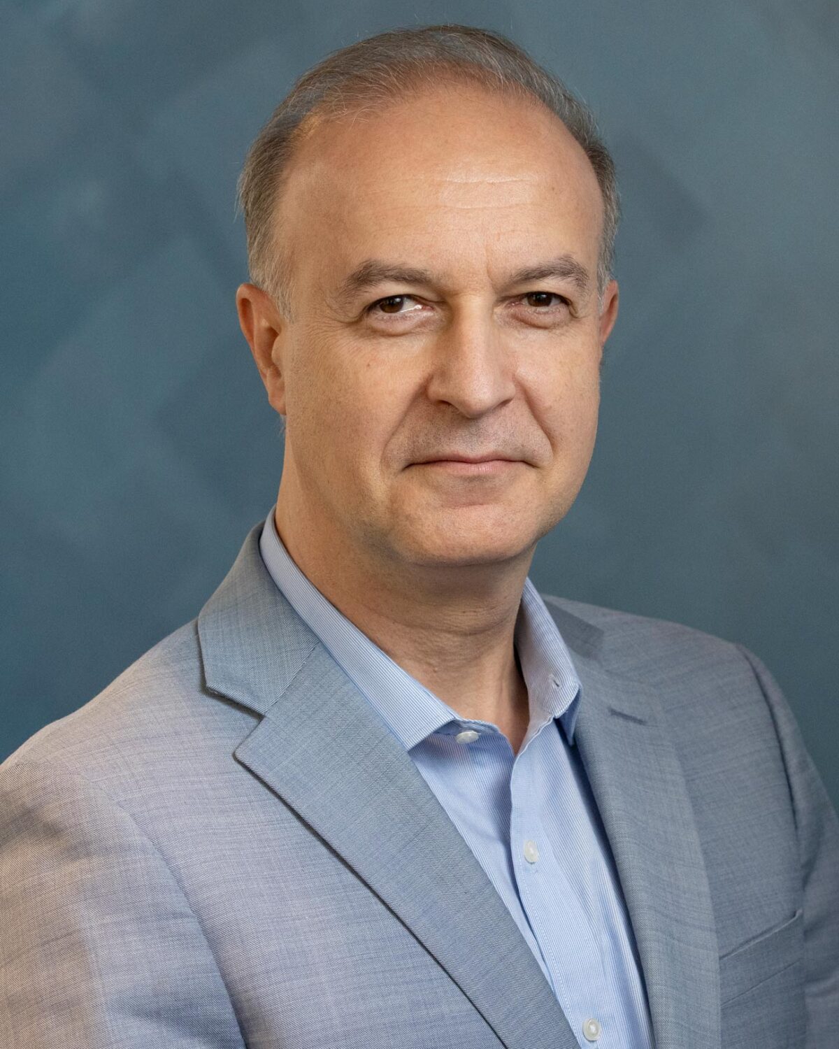 A middle-aged man with short hair wearing a light blue dress shirt and a grey blazer.