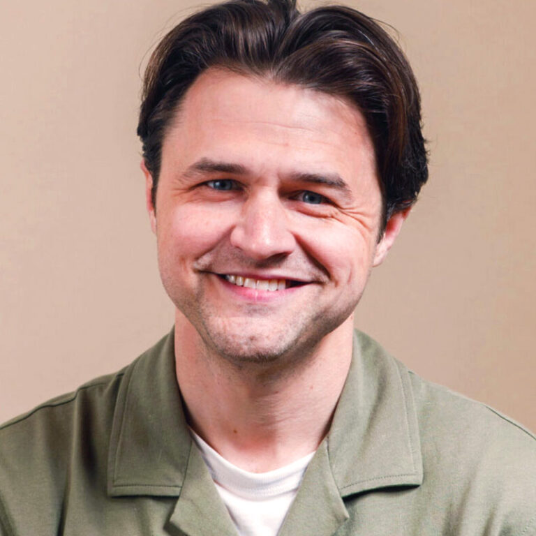 A man with dark hair smiling at the camera, wearing an olive green collared shirt over a white t-shirt.