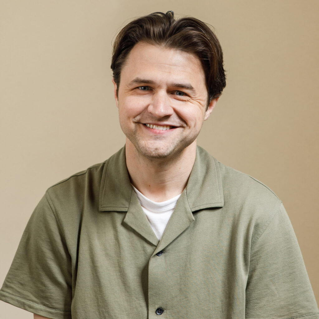 A man with short brown hair, wearing a green button-up shirt over a white t-shirt, smiles while sitting in front of a plain beige background.