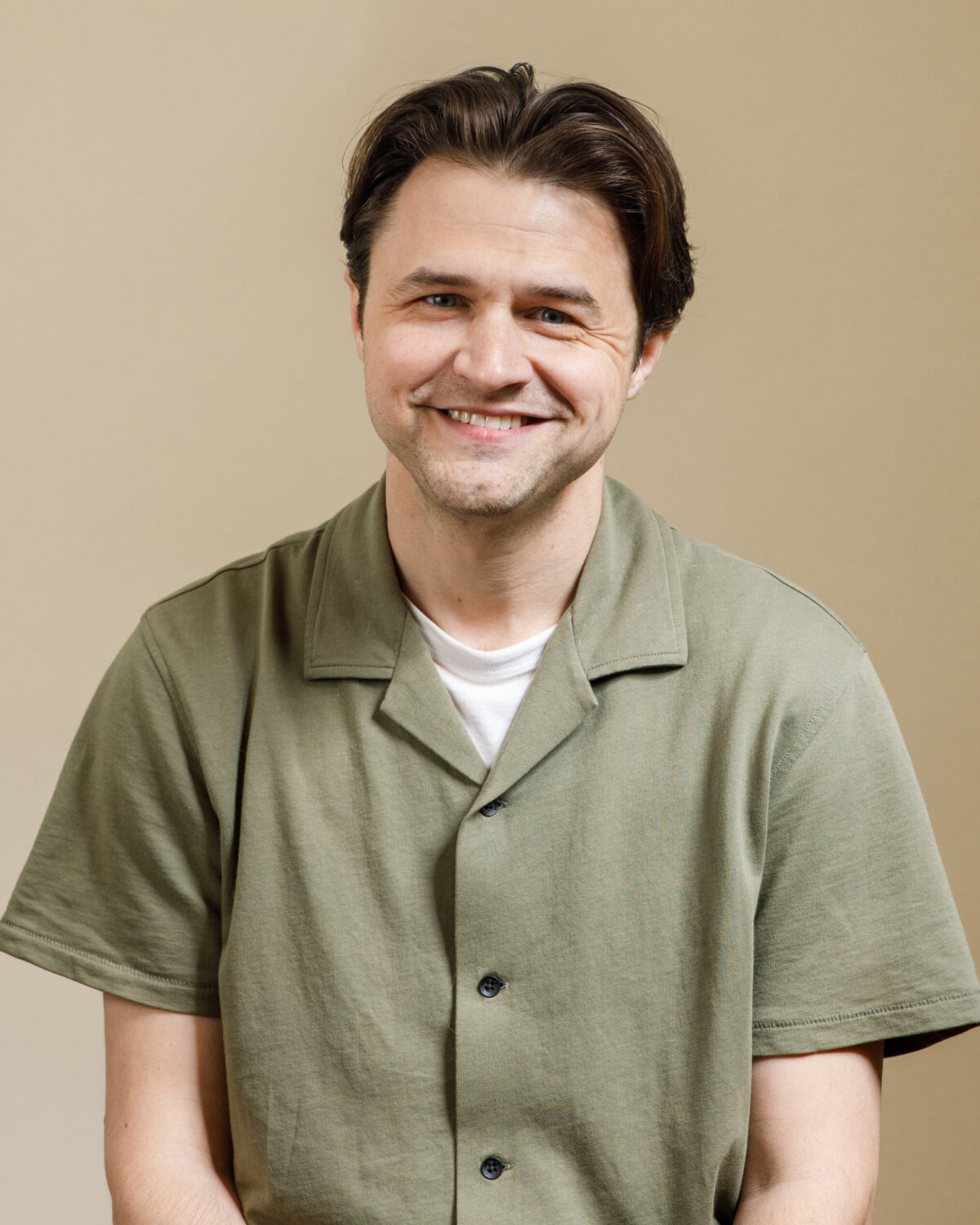 A man with short brown hair, wearing a green button-up shirt over a white t-shirt, smiles while sitting in front of a plain beige background.