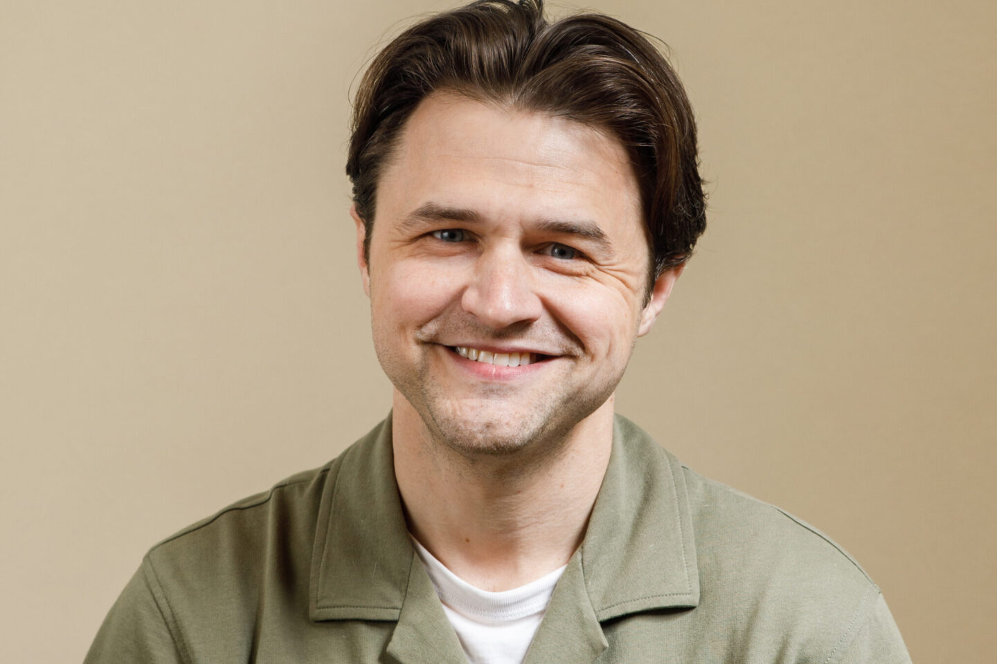 A man with short brown hair, wearing a green button-up shirt over a white t-shirt, smiles while sitting in front of a plain beige background.