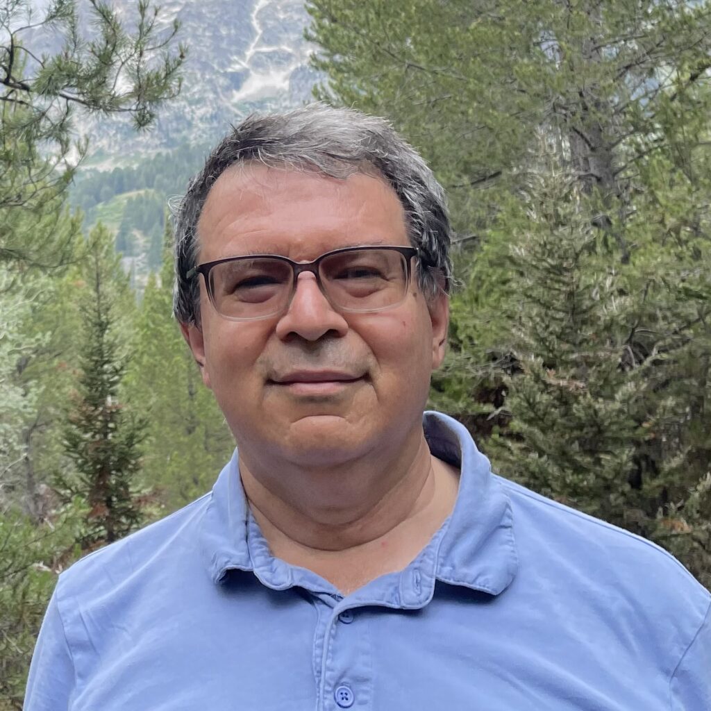 A man wearing glasses and a blue polo shirt stands outdoors in a forest with pine trees, with mountains visible in the background under a cloudy sky.