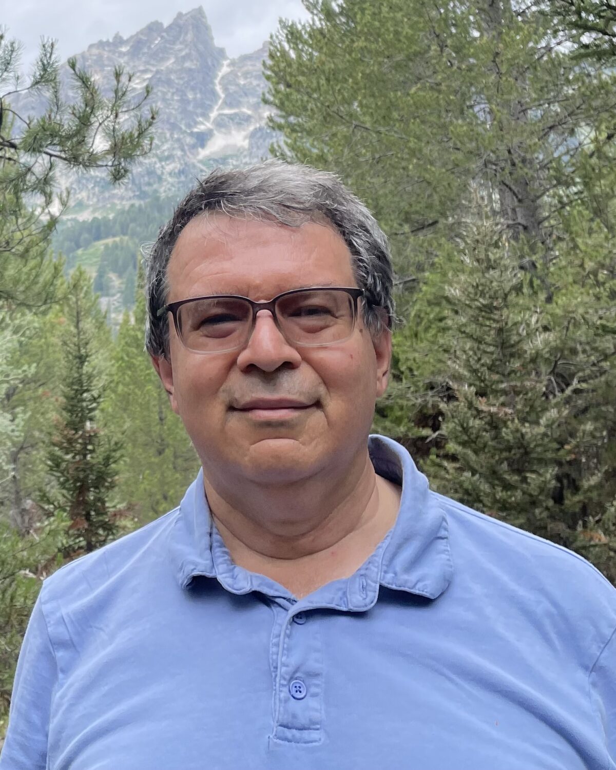 A man wearing glasses and a blue polo shirt stands outdoors in a forest with pine trees, with mountains visible in the background under a cloudy sky.