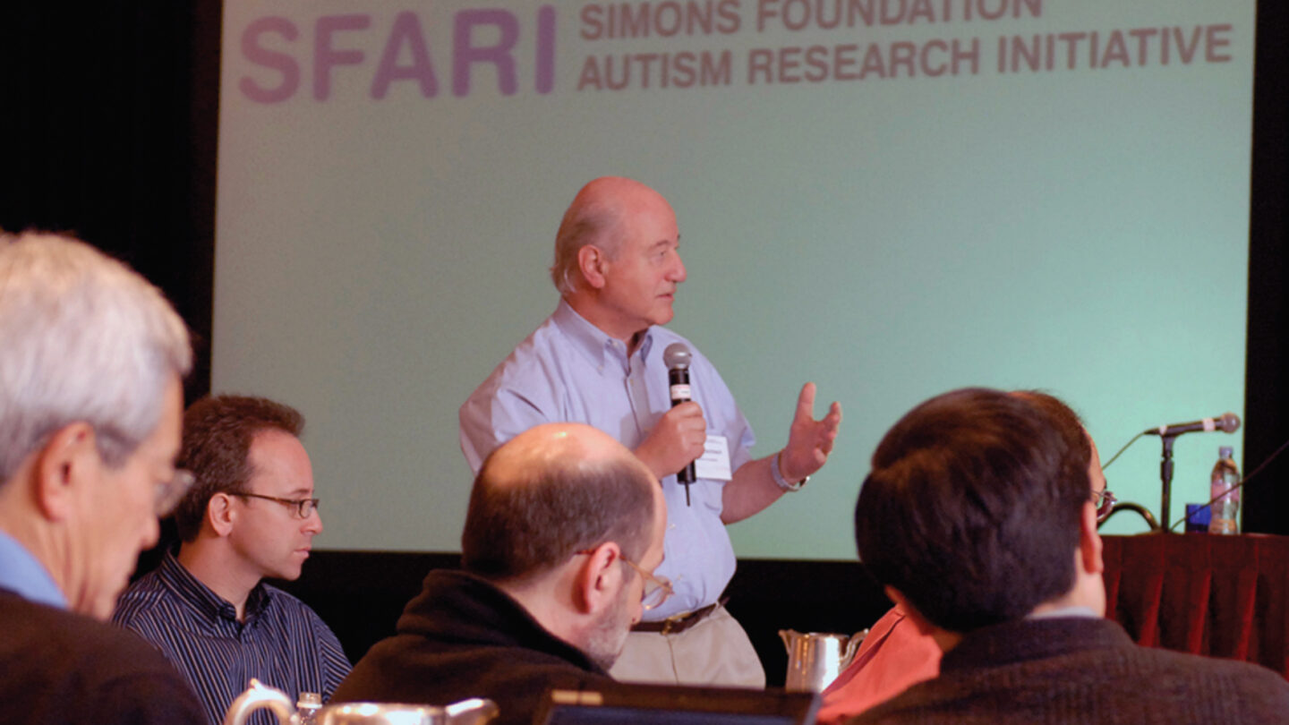 A man speaks into a microphone at a podium in front of a screen displaying the Simons Foundation Autism Research Initiative (SFARI) logo.