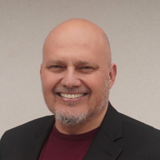 A bald man with a short beard is smiling, wearing a dark blazer over a maroon shirt, standing in front of a plain light-colored background.