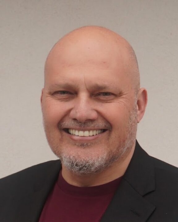 A bald man with a short beard is smiling, wearing a dark blazer over a maroon shirt, standing in front of a plain light-colored background.