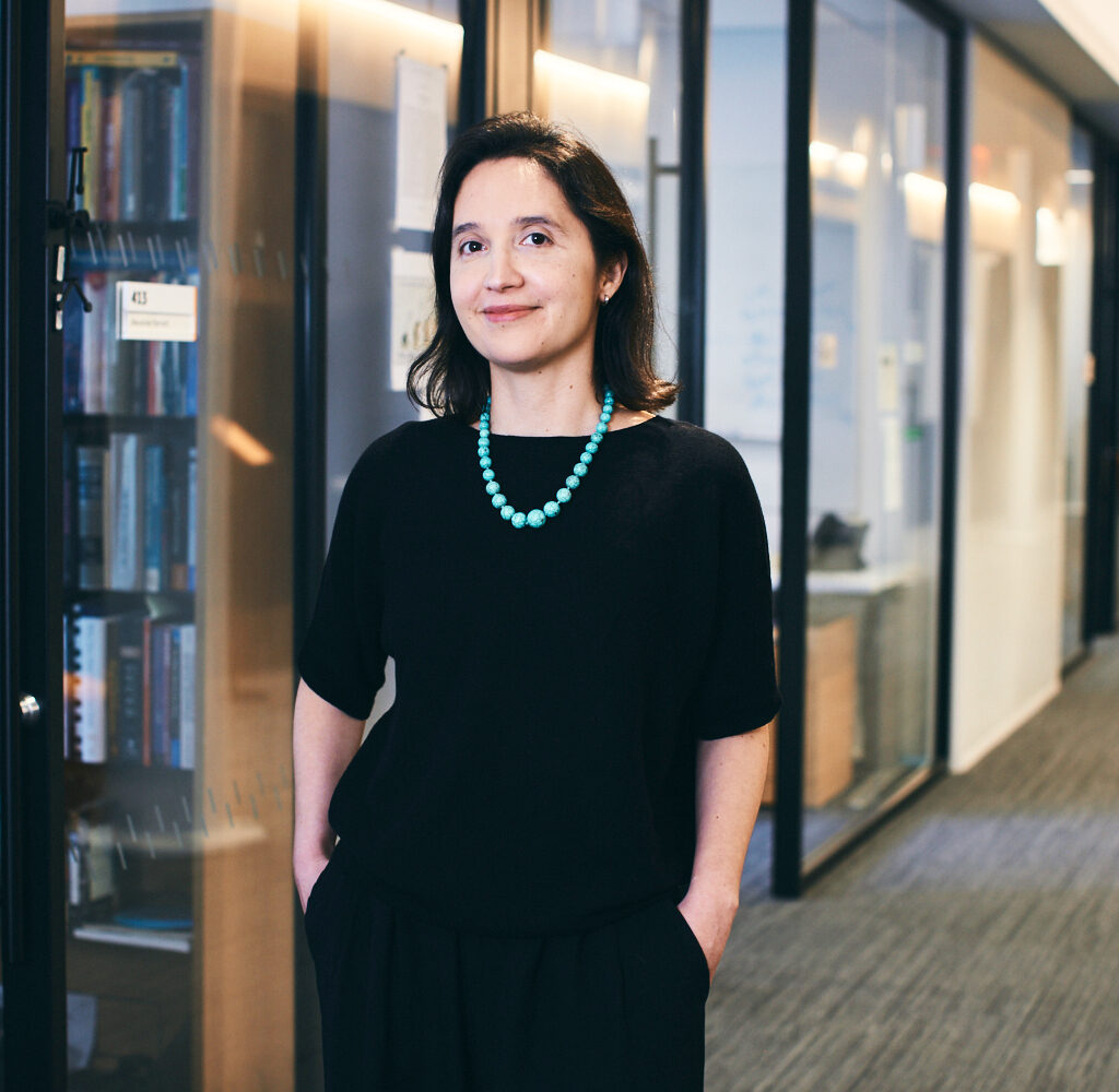 A woman with dark hair, wearing a black outfit and turquoise necklace, stands with hands in pockets in a modern office hallway lined with glass walls and bookshelves.