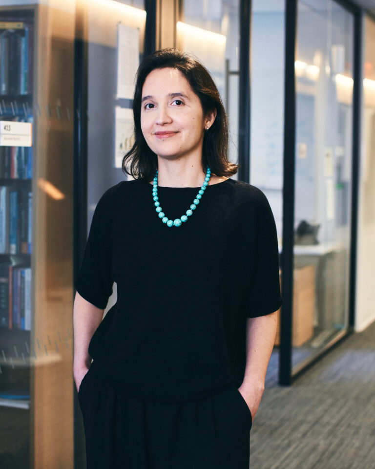 A woman with dark hair, wearing a black outfit and turquoise necklace, stands with hands in pockets in a modern office hallway lined with glass walls and bookshelves.
