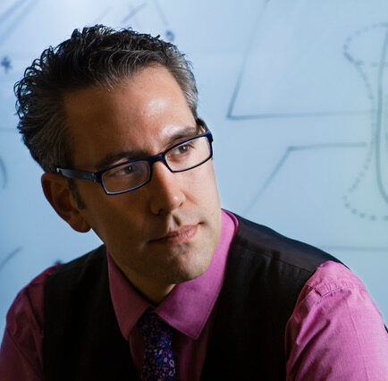 A person wearing glasses, a pink shirt, and a vest looks thoughtfully to the side. Behind them is a whiteboard with abstract diagrams and arrows drawn on it.