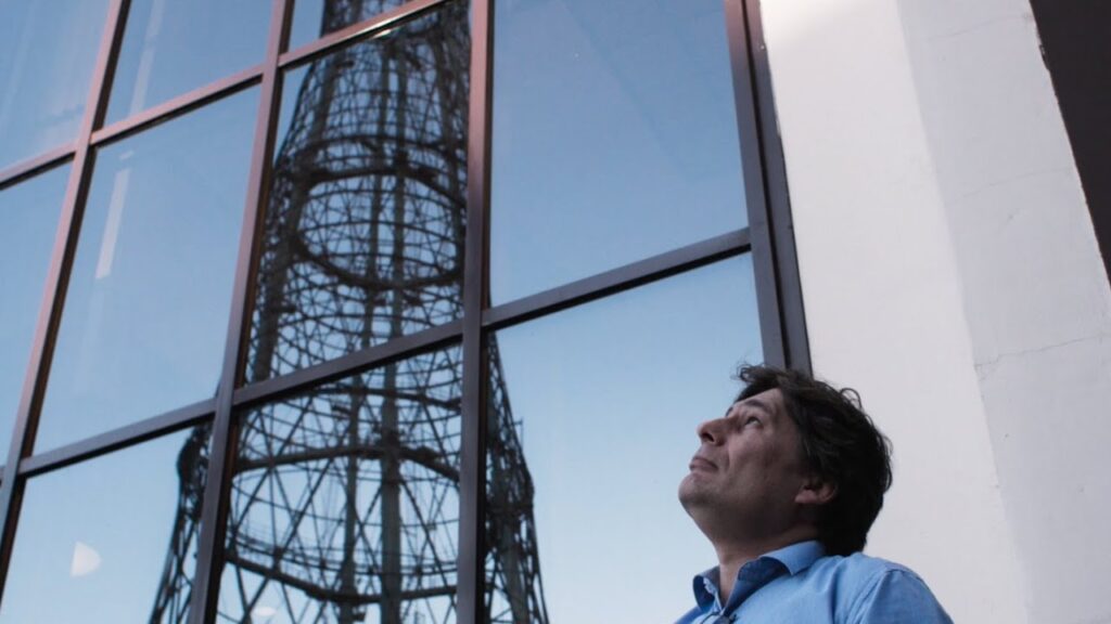 A man looking up at the reflection of the Shukhov Tower in a large glass building window.