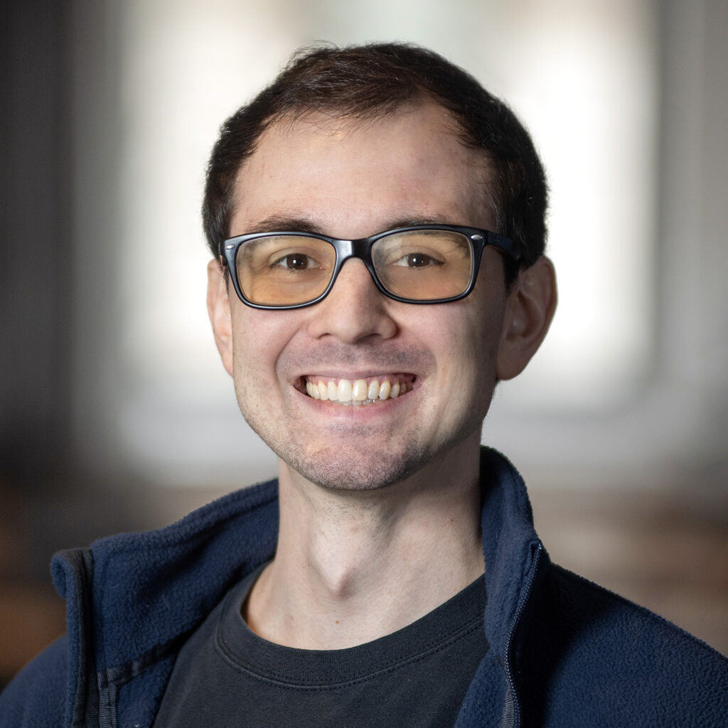 A young man wearing glasses and a dark jacket smiles at the camera. The background is softly blurred, focusing attention on his face.