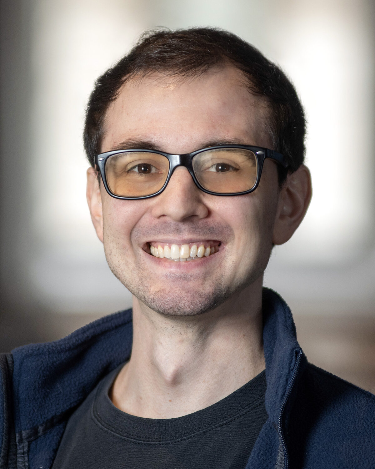 A young man wearing glasses and a dark jacket smiles at the camera. The background is softly blurred, focusing attention on his face.