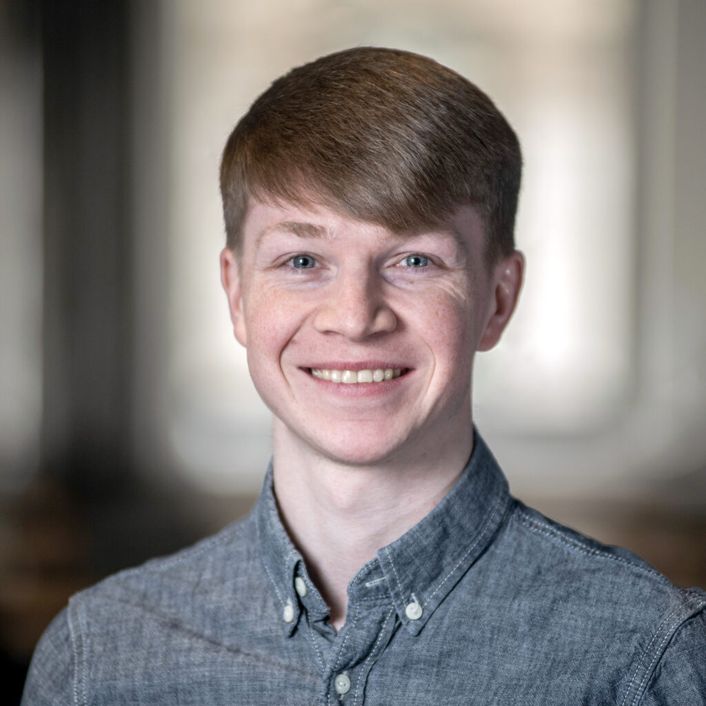 A young person with short light brown hair, wearing a button-up gray shirt, is smiling at the camera. The background is softly blurred, suggesting an indoor setting with natural light coming through windows.