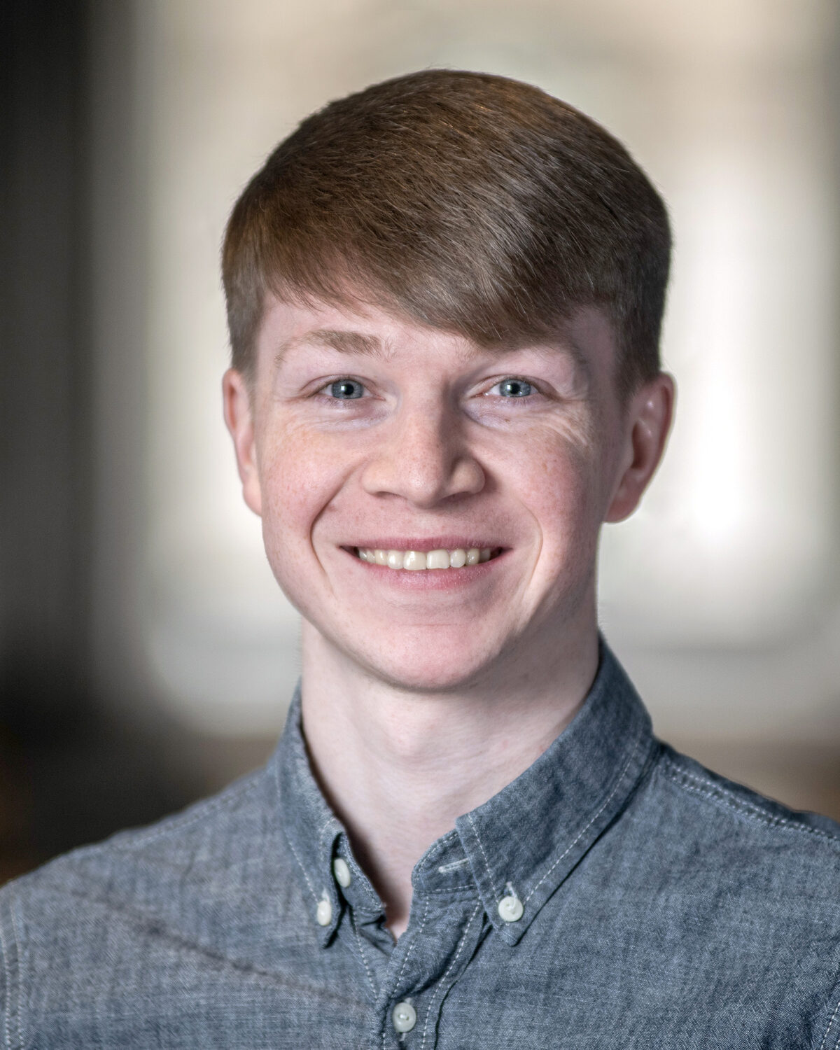 A young person with short light brown hair, wearing a button-up gray shirt, is smiling at the camera. The background is softly blurred, suggesting an indoor setting with natural light coming through windows.