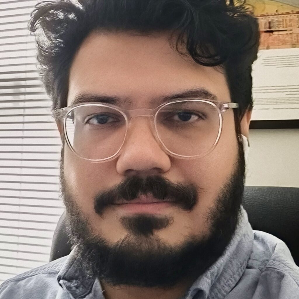 A person with dark hair, beard, and mustache is wearing clear glasses and a light blue shirt, sitting in an office with blinds and a framed poster in the background.