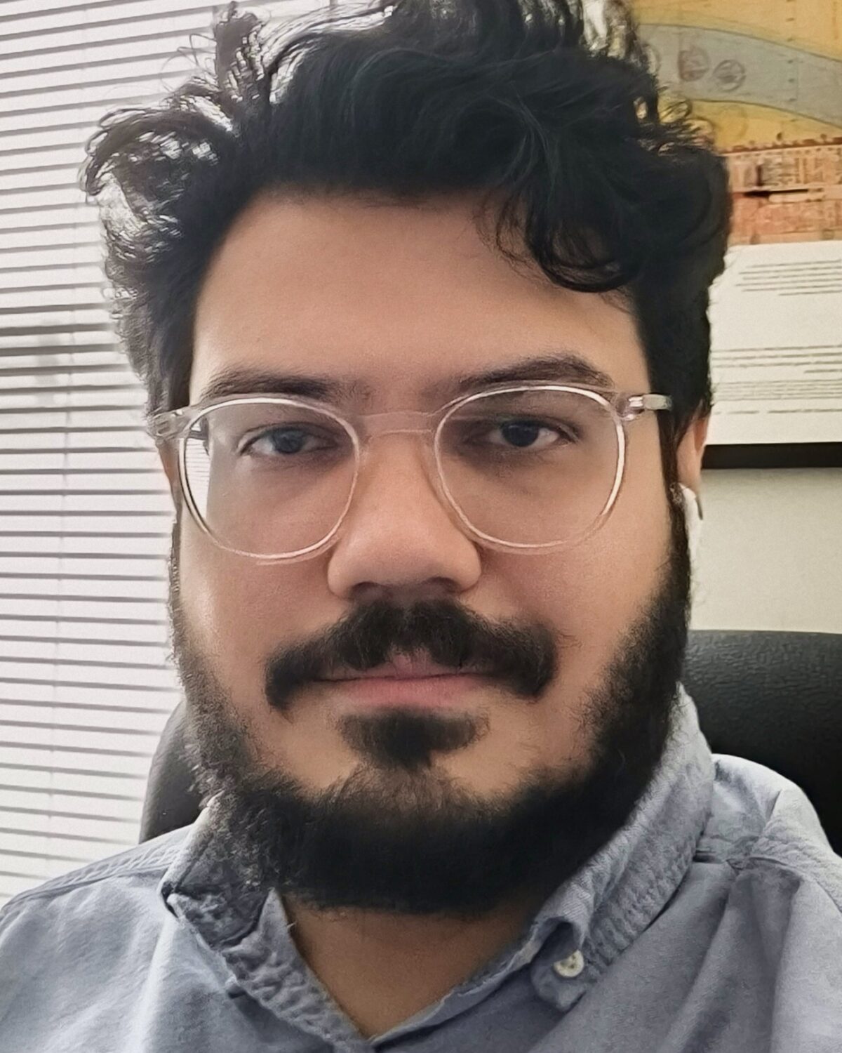 A person with dark hair, beard, and mustache is wearing clear glasses and a light blue shirt, sitting in an office with blinds and a framed poster in the background.