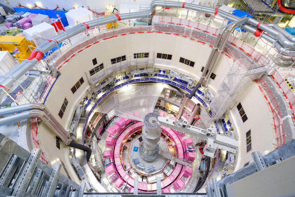 A large circular construction site with multiple floors, scaffolding, and machinery, viewed from above. The central area contains pink-covered structures and metal framework, surrounded by cables and industrial equipment.